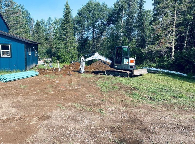 A small excavator digs near a blue house, piles of dirt and pipes on a grassy lot.