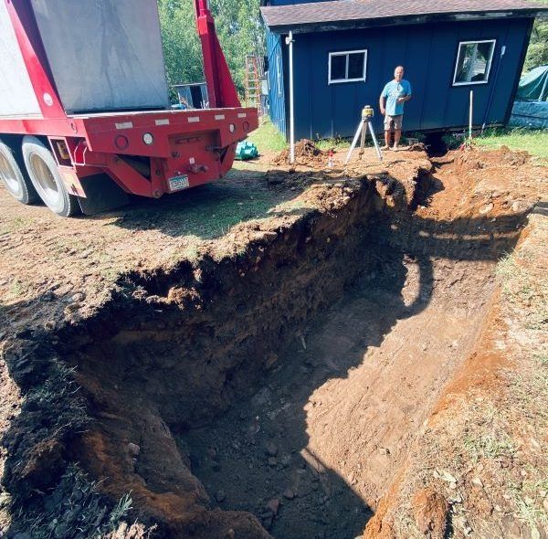 Man watches long trench excavation beside a blue building; red truck in background.
