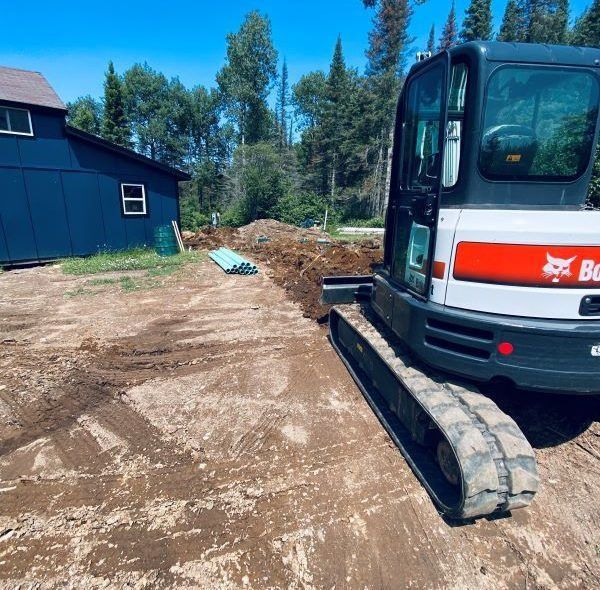 Bobcat excavator next to a blue building, working on a dirt construction site on a sunny day.