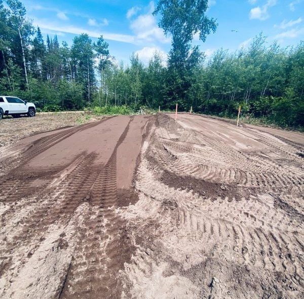 Dirt track with tire tracks and a truck, set against a forest and blue sky.