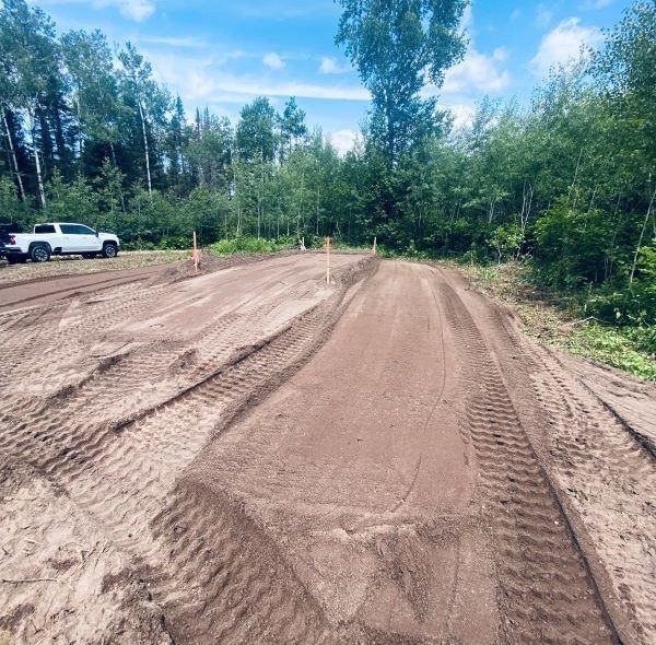 Dirt track with tire tracks, trees in the background, and a white truck parked to the left.