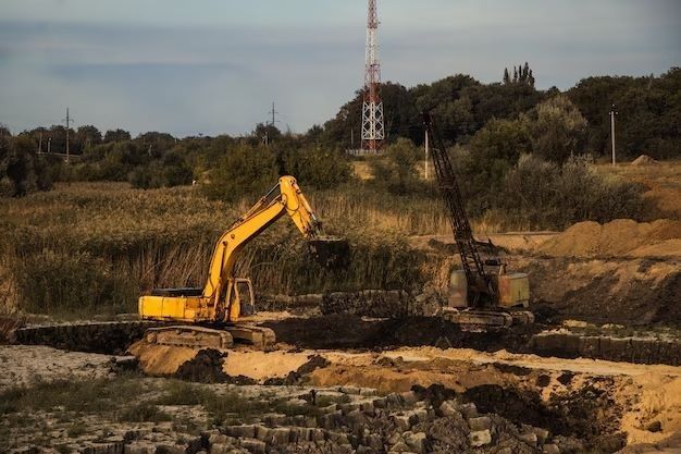 Yellow excavator and crane digging in a quarry, with a radio tower in the background.