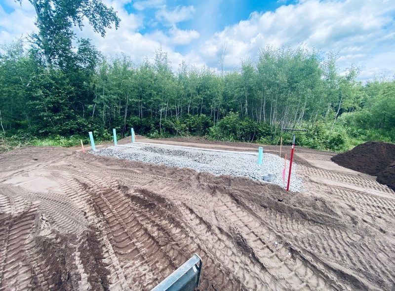 Gravel-lined trench with vertical pipes in a dirt field; trees in the background under a blue sky.