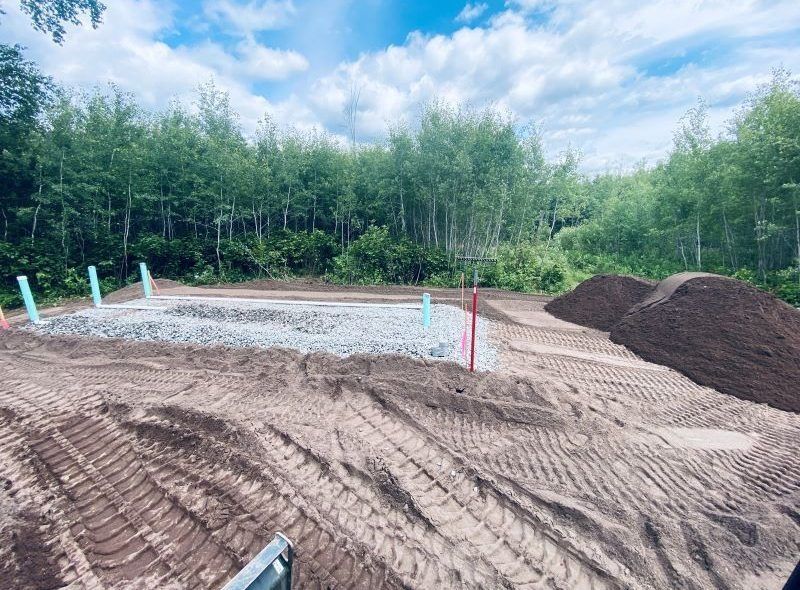 Construction site: gravel foundation, soil piles, blue pipes, brown earth, and trees under a cloudy sky.
