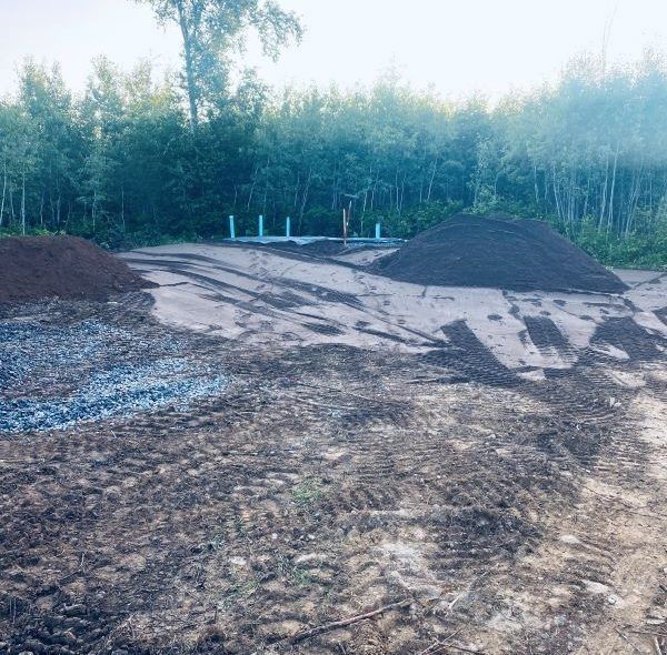A cleared construction site with piles of dirt and gravel, with a forest backdrop.