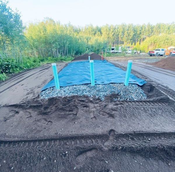 Construction site with gravel and three upright blue pipes in the center, covered with a blue tarp.
