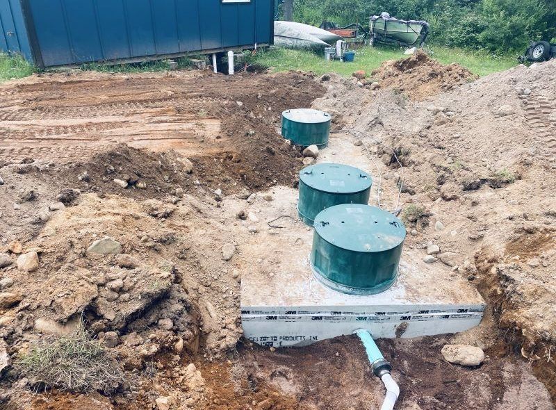 Installation of a septic system; three green tanks in a trench, next to a building, outdoor setting.