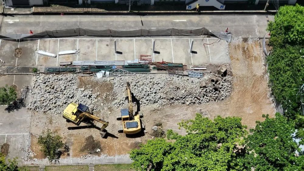 Two excavators on a construction site with demolished concrete, a parking lot, and green trees overhead.
