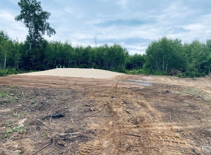Dirt road leading to a cleared sandy area surrounded by trees under a cloudy sky.