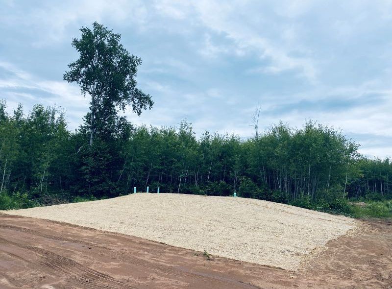 A gravel-covered RV site with utility hookups, surrounded by trees, under a cloudy sky.