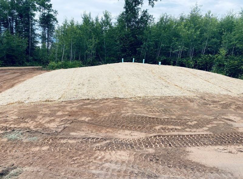 A large pile of light-colored gravel with white pipes on top, surrounded by dirt, against a treeline.
