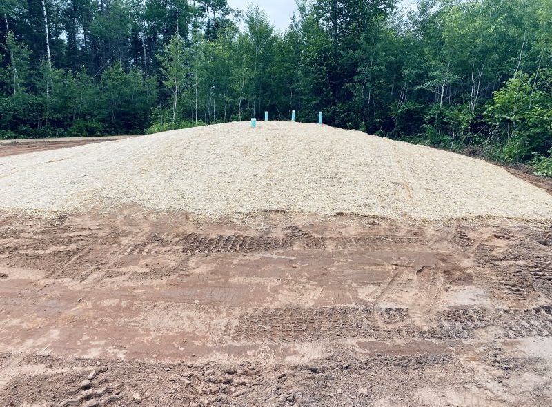 A gravel mound with blue pipes, surrounded by dirt and trees in a wooded area.