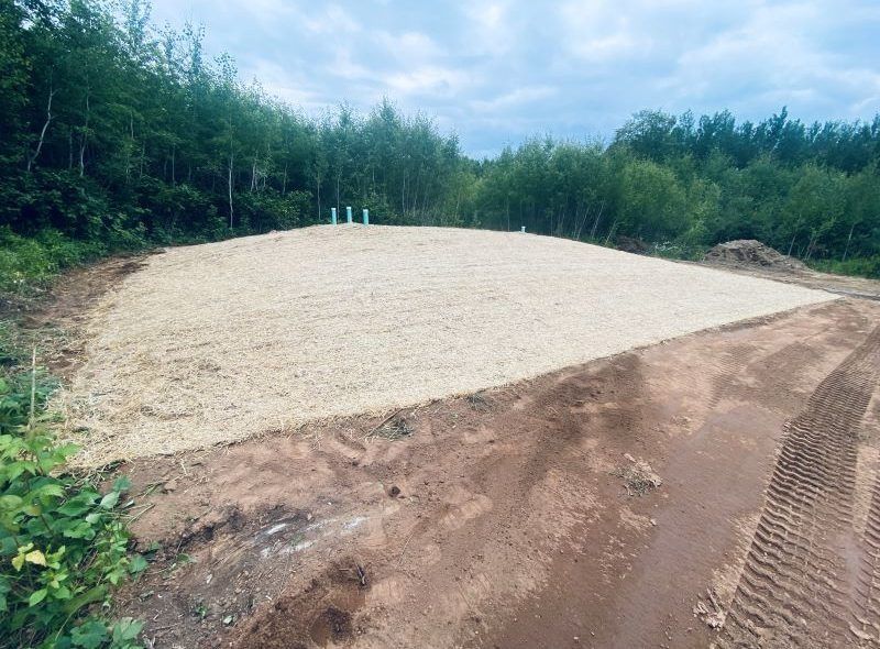 Gravel bed in a clearing, with white pipes emerging. Forest background. Overcast sky.