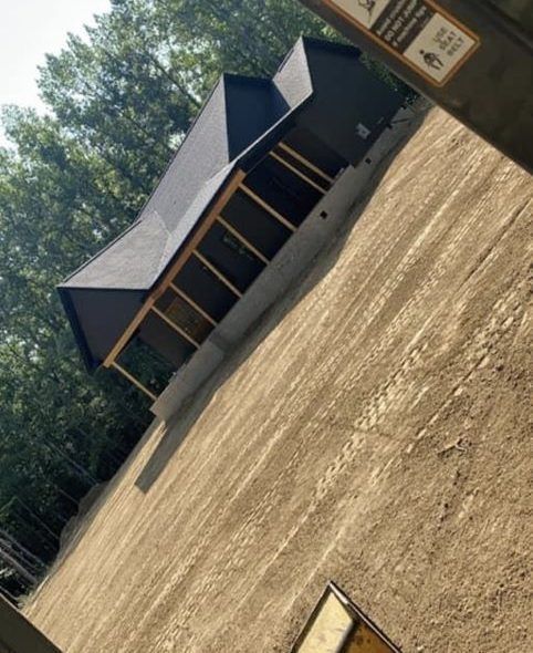 A small, dark-roofed building with a screened porch sits on a graded dirt surface with trees in the background.