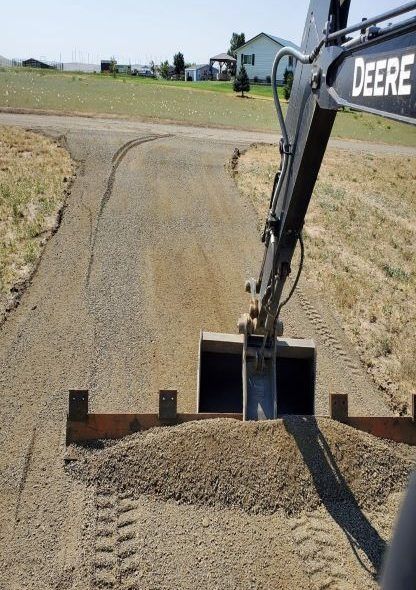 Gravel road being graded by a Deere backhoe. Brown gravel and dirt. Sunny day.