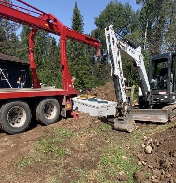 A red boom truck lifts a concrete septic tank near a Bobcat excavator on a grassy construction site.