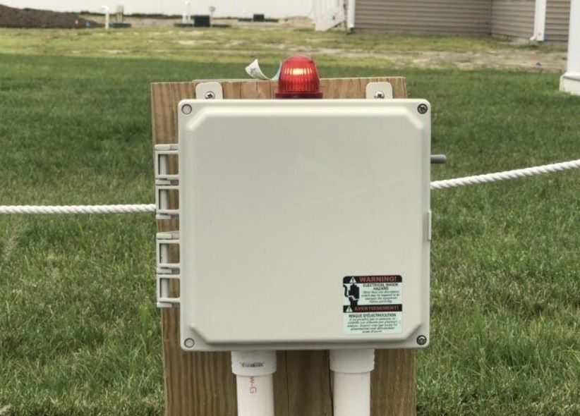 White electrical box with a flashing red light attached to a wooden post in a grassy area.