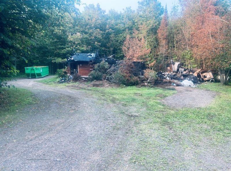 Gravel driveway leads to a small log cabin nestled amongst trees and brush. A green dumpster sits to the left.