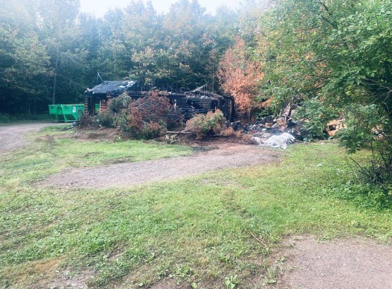 Burnt-out building surrounded by charred trees, bushes, and grass, set in a green, wooded area.