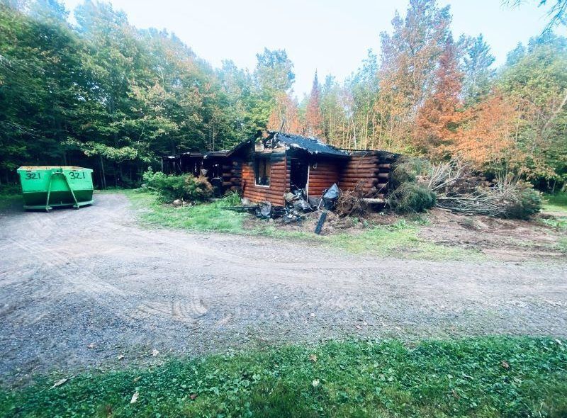 Burned-out log cabin with debris, green dumpster, gravel driveway, surrounded by fall foliage and trees.