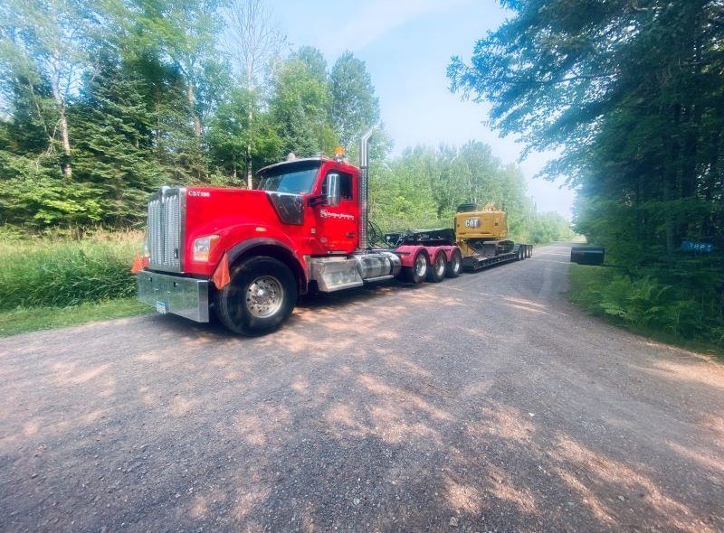 Red semi-truck with heavy equipment on a trailer, parked on a gravel road surrounded by trees.
