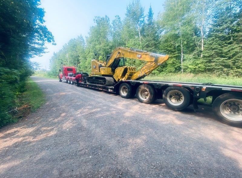 A red semi-truck hauling a yellow excavator on a long flatbed trailer on a gravel road, surrounded by trees.