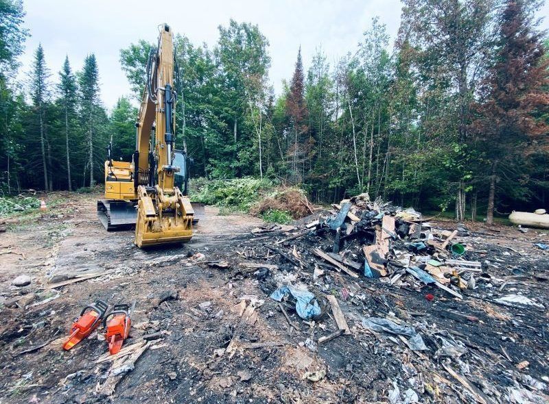 Yellow excavator clearing debris in a wooded area. Two chainsaws lie on the ground.