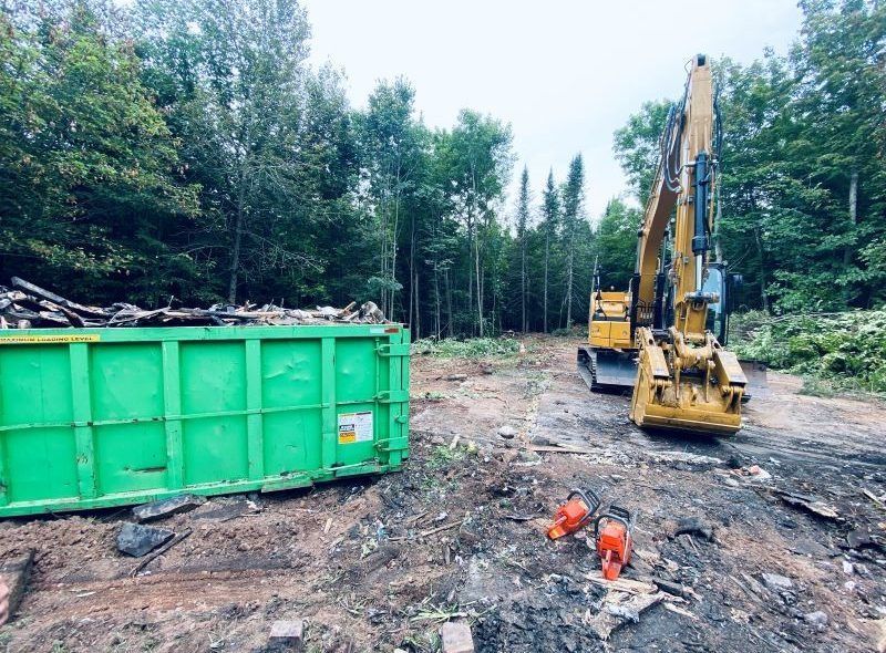 Green dumpster and yellow excavator on a cleared patch of land in a forest, surrounded by trees.