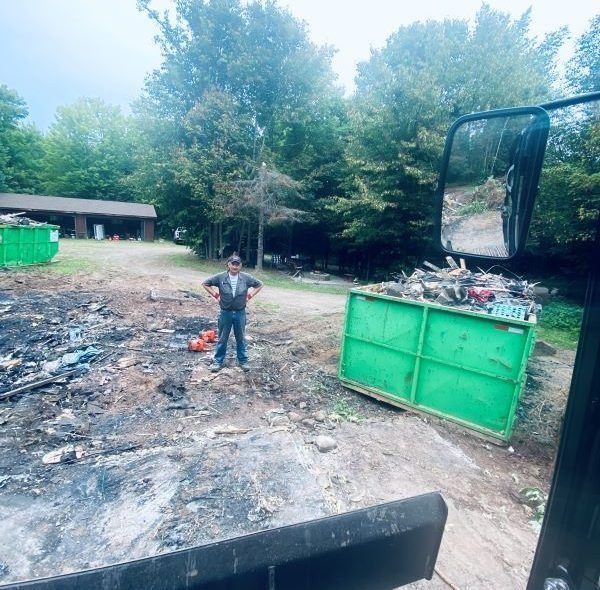 Man standing near green dumpster filled with wood, trees in the background, and a building.