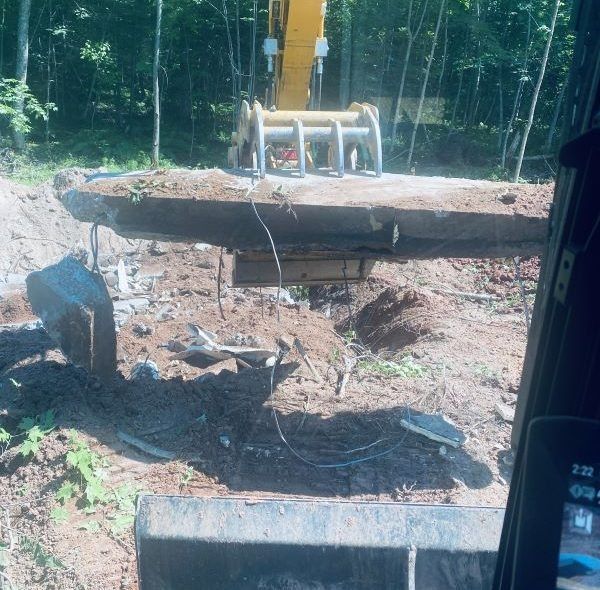 Excavator lifting concrete slab in a dirt clearing, surrounded by trees.
