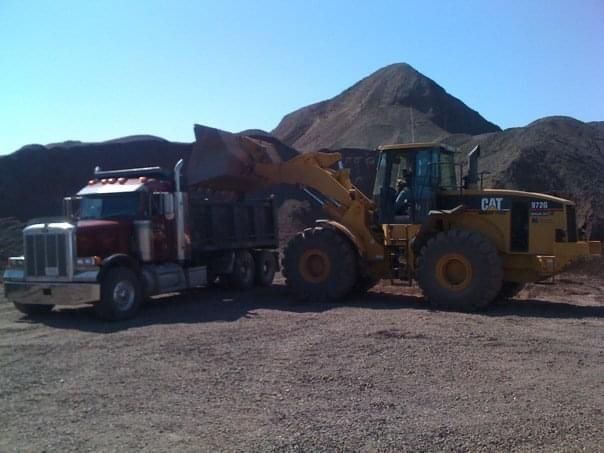 A red semi-truck is loaded with gravel by a yellow Caterpillar front-end loader against a mountain of gravel.