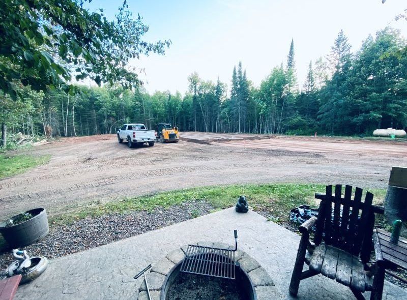 Gravel clearing with truck and excavator. Fire pit in foreground. Wooded area in background.