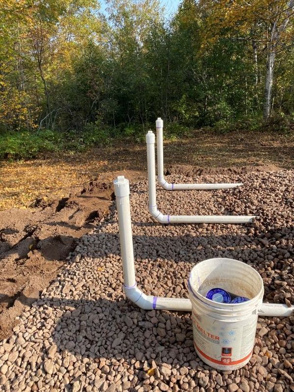 Pipes protruding from gravel ground, connected to a bucket. Outdoor setting with trees in background.