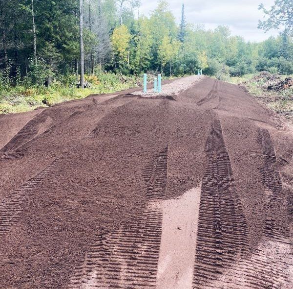 Pile of dark soil with tire tracks in a wooded area, with blue pipes emerging.