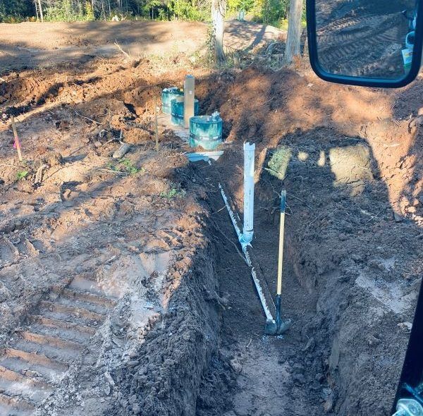 Trench dug in dirt with pipes, tools, and blue containers. Daytime outdoor setting.
