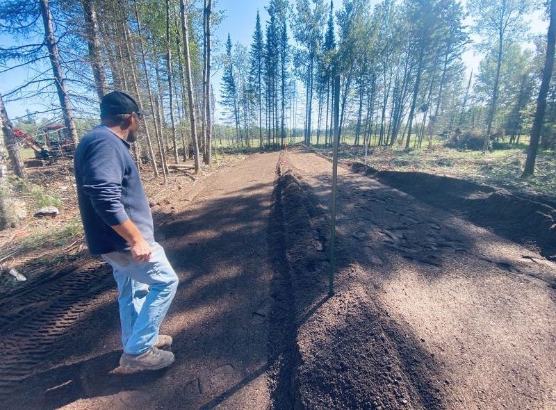 Man in jeans and blue shirt surveys freshly graded dirt trail in a forest.