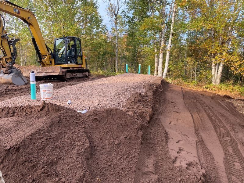 Construction site with an excavator, gravel pile, and dirt in a wooded area.