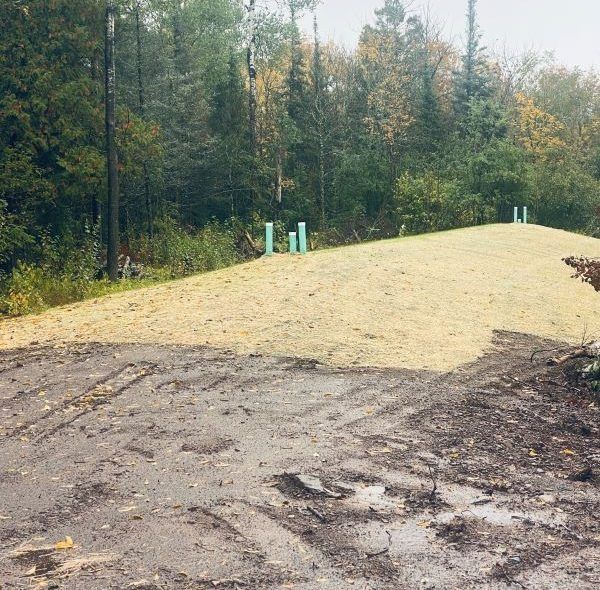 A dirt road leads to a grassy hill with green pipes, surrounded by a forest.
