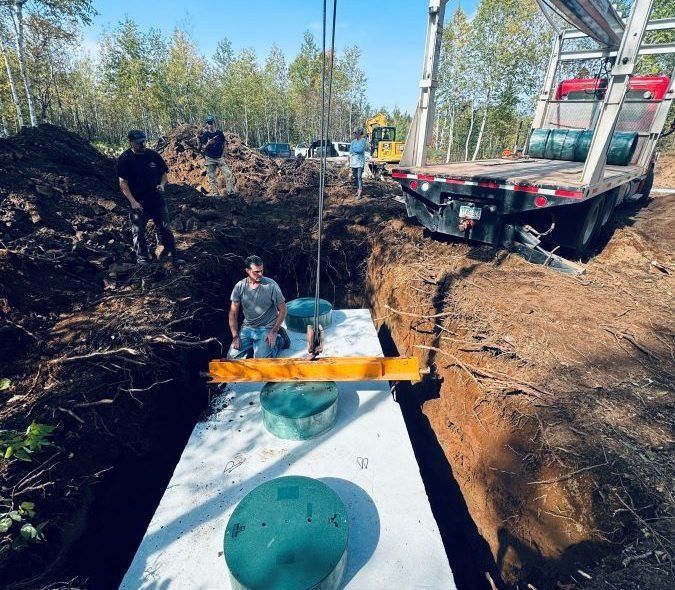 Workers install a septic tank into a trench with a crane in a wooded area.