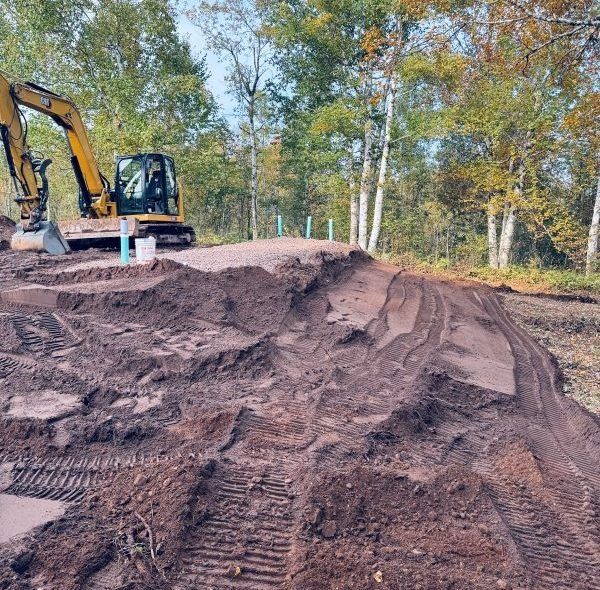 A yellow excavator shapes a dirt bike track in a wooded area.
