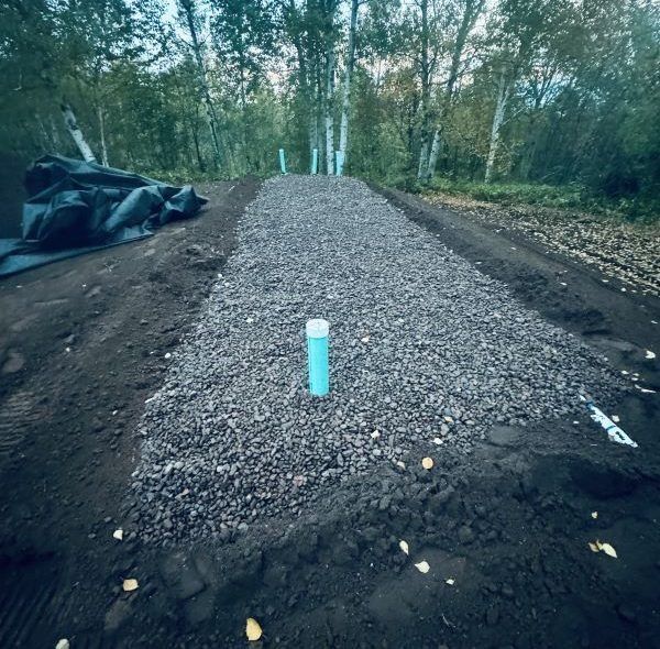 Gravel path with blue PVC pipe, surrounded by dirt and trees.