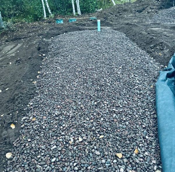 Gravel-filled trench with a blue pipe, likely for drainage, outdoors. Dark soil and trees in the background.