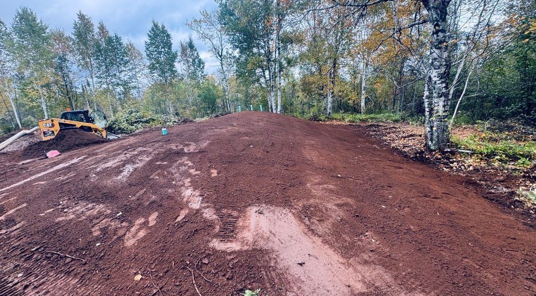 Brown mound of dirt in a wooded area, a bulldozer is on a nearby hill.