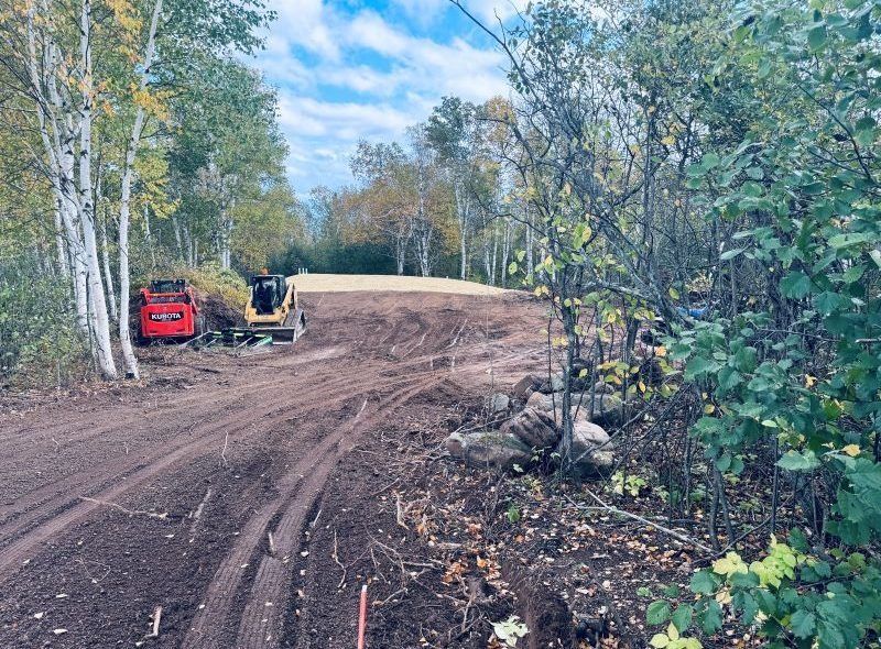 Dirt road construction site with heavy machinery and trees under a blue sky.
