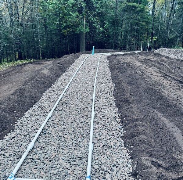 Gravel-filled trench with parallel white pipes in a wooded area, likely for a septic system.