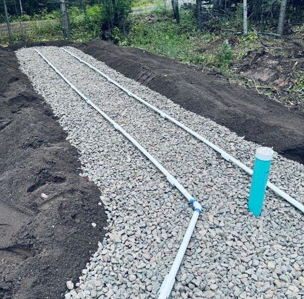 Gravel-filled trench with white pipes and a blue vent pipe; part of a septic system being installed.