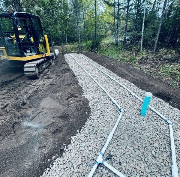 Excavator and gravel bed with PVC pipes, likely for septic system installation in a wooded area.