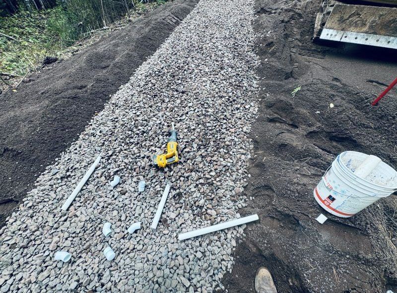 Gravel-filled trench with PVC pipes, tool, and bucket on a hillside.