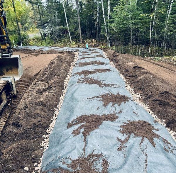 A long, dark fabric-covered area is being prepped for planting, with a backhoe and forest backdrop.