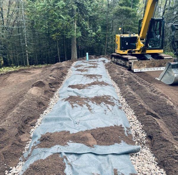 An excavator working on a long trench lined with gravel and fabric, dirt piled on top. Forest in background.